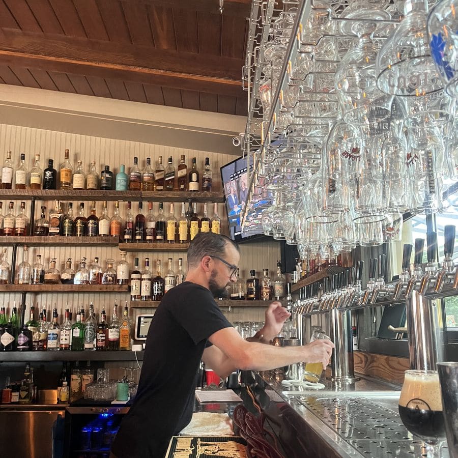 A bartender pulling a pour at the tap wall, bourbon shelves behind him